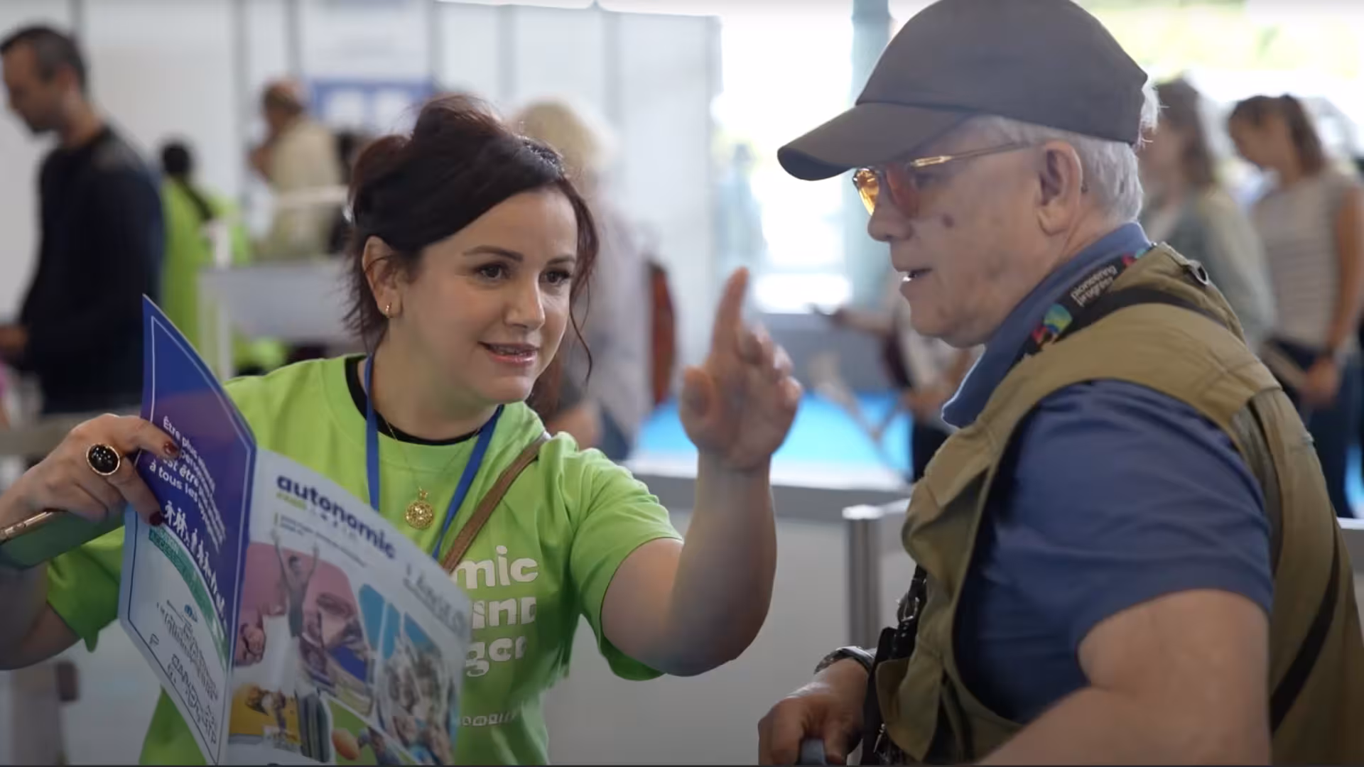 Une exposante portant un t-shirt vert Autonomic échange avec un visiteur et lui indique une direction sur le salon, tout en tenant un dépliant d’information.